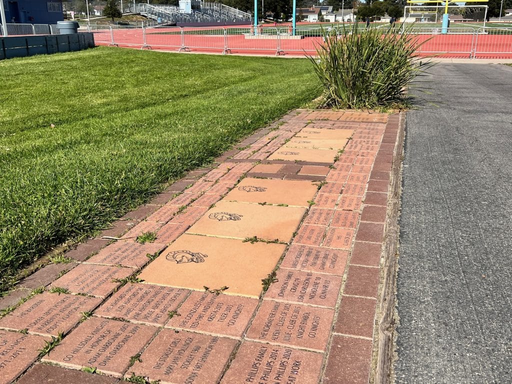 Engraved bricks line a walkway next to grass, near a sports track and field.